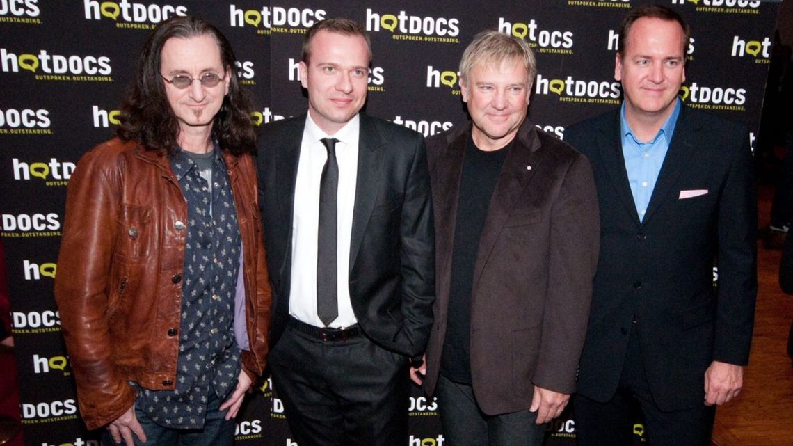 Three members of Rush band stand next to Hot Docs' Chris McDonald in front of a Hot Docs logo backdrop.