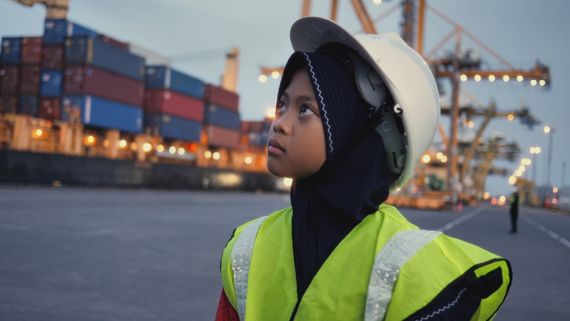 a young girl in a yellow construction vest and hard hat with a boat full of shipping containers in the background